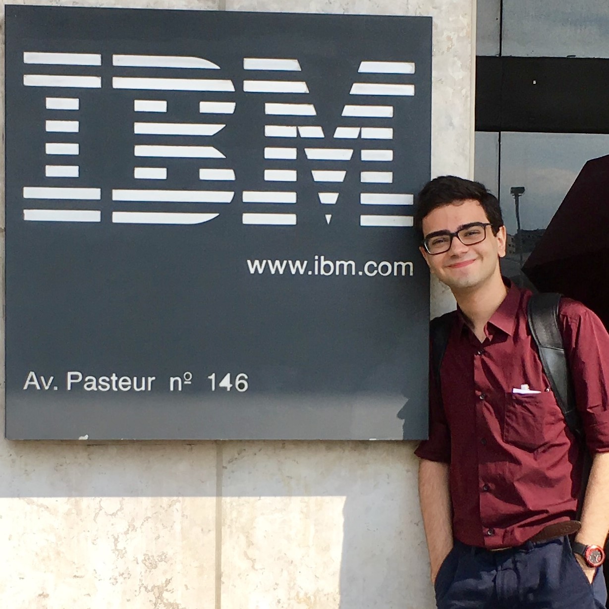 Foto minha em frente ao prédio da IBM no Rio de Janeiro. Ao meu lado está uma placa quadrada, com o logotipo da IBM brando, num fundo preto.